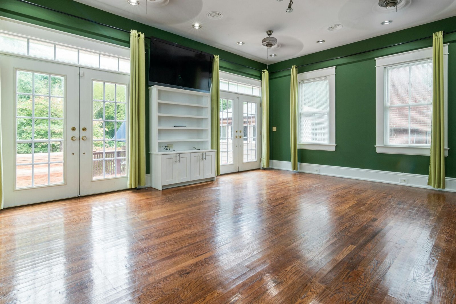 Empty sunlit room with hardwood floor, large windows, and green walls.